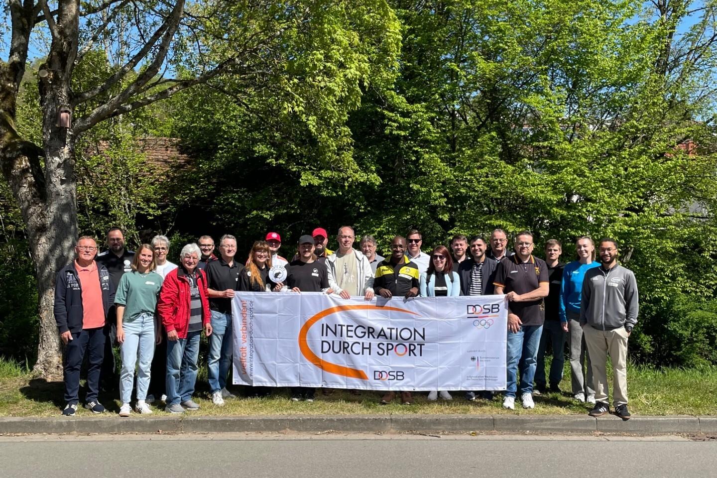 Gruppenschäffungsfoto mit Banner „Integration durch Sport“ und vielen Teilnehmern vor grüner Kulisse.
