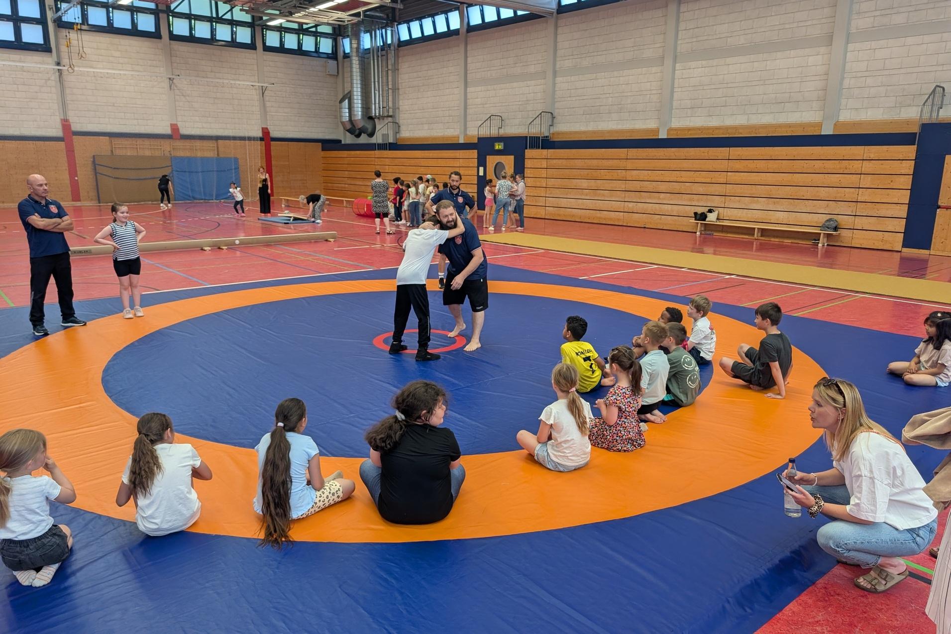 Zwei Kinder üben Ringen auf einer Matte, umgeben von beobachtenden Kindern und Trainern in einer Sporthalle.