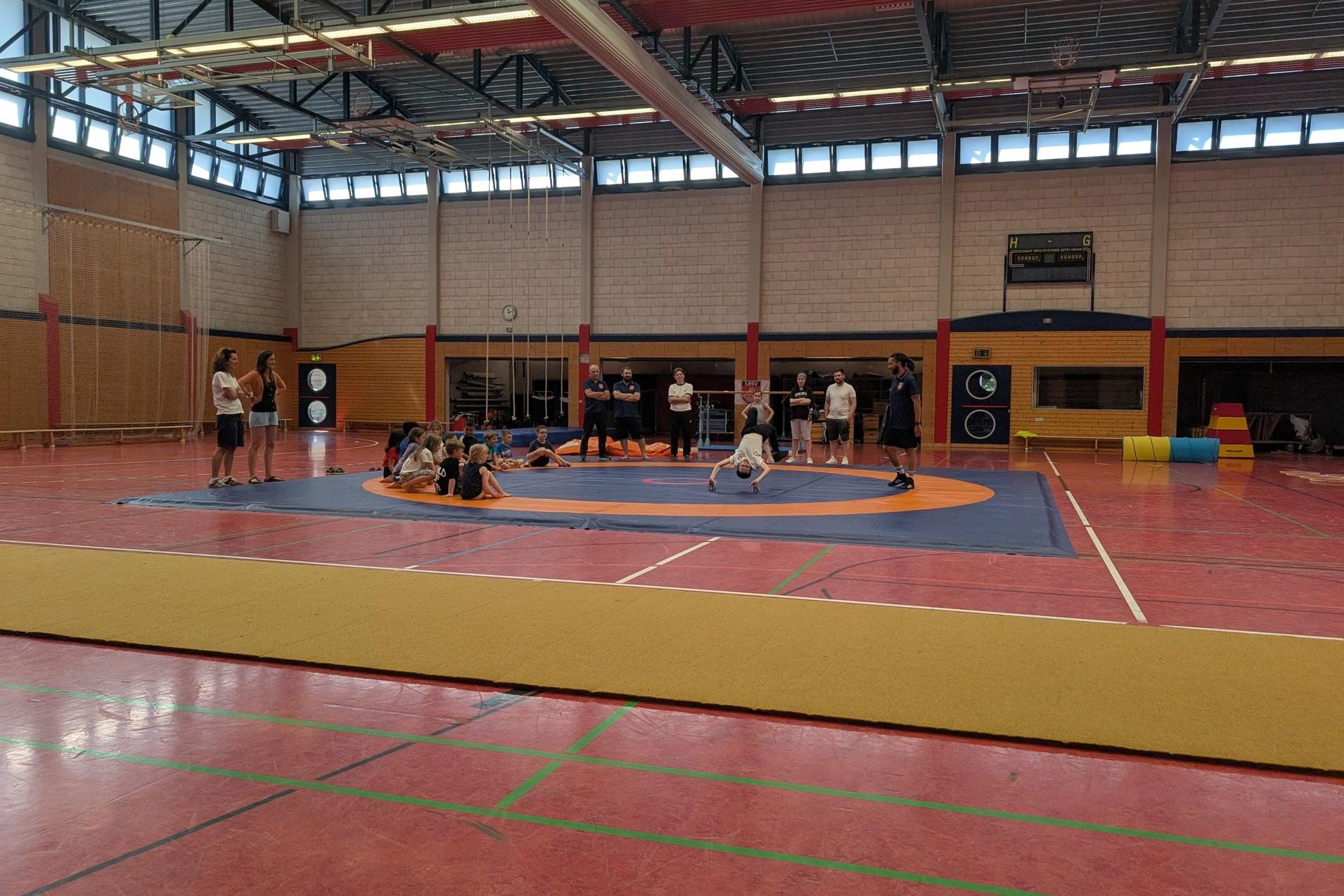 Mehrere Personen trainieren auf einer großen, orange-blauen Sportmatte in einer hellen Halle.